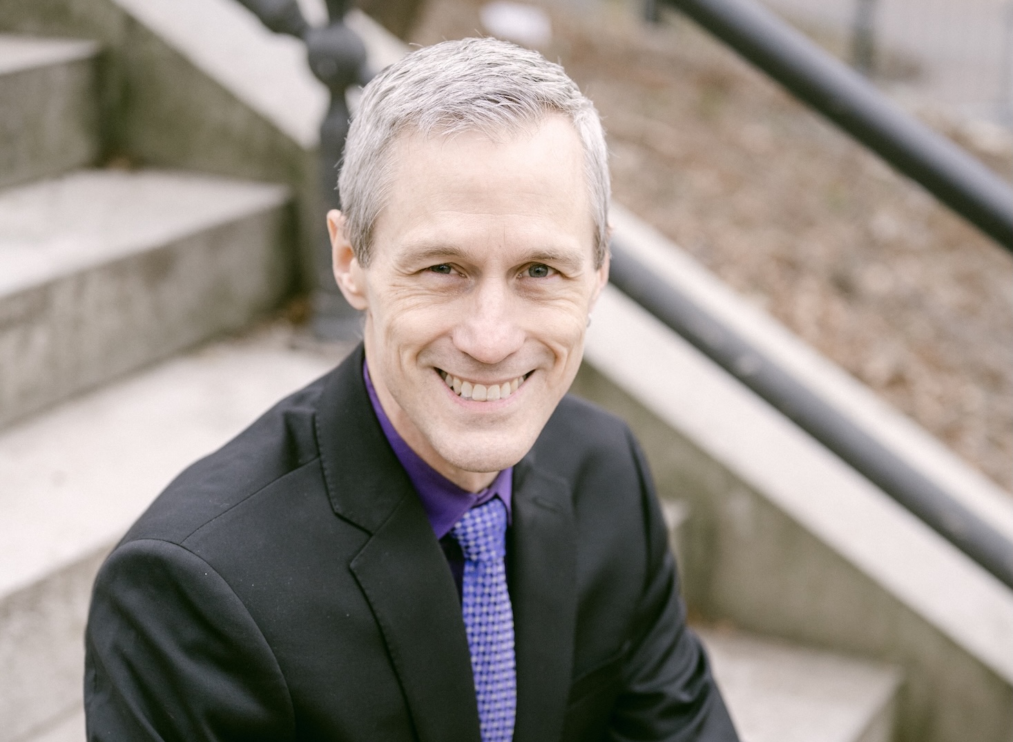 Ross Murray sits on outdoor steps wearing a black suit jacket, black shirt, and purple tie.