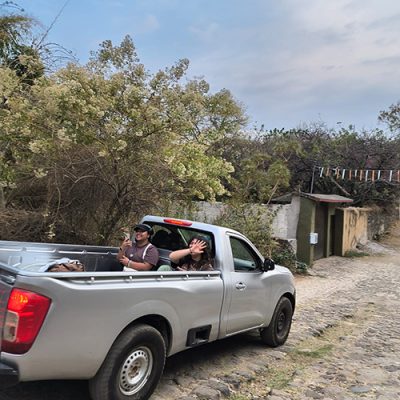 f68876d4-ce19-4610-acdb-6e6ec496e20d Two people smiling and riding in the back of a silver pickup truck along a cobblestone road
