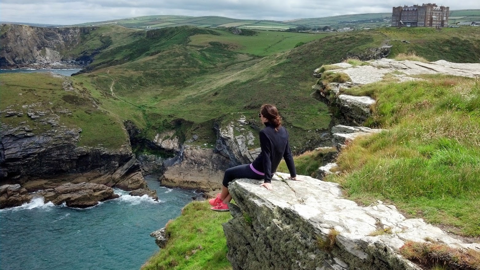 A woman sits atop a cliff in Cornwall, England. She is looking out over the ocean and the rolling hills of the English countryside. At this site she is experiencing the natural elements of the wind in her face, the ocean mist, the solid stone of the cliff, and the warmth of the sun. –photo credit K.McHale