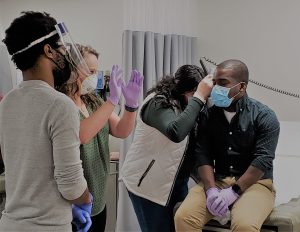 Four masked people, two observing another checking ears