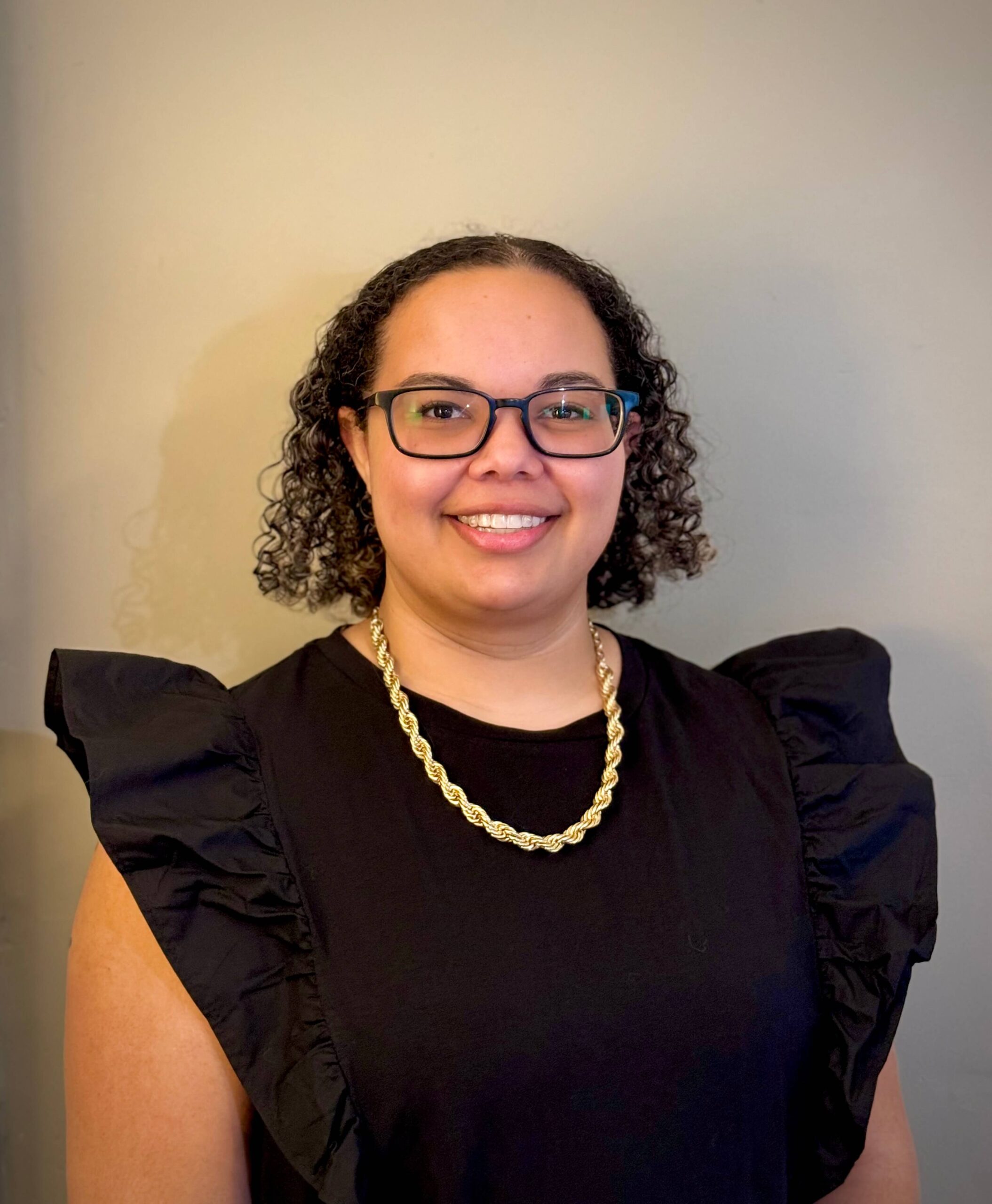 bust up photo of Miranda in a black top that has frilly sleeves. She is smiling and wearing dark-rimmed glasses. 