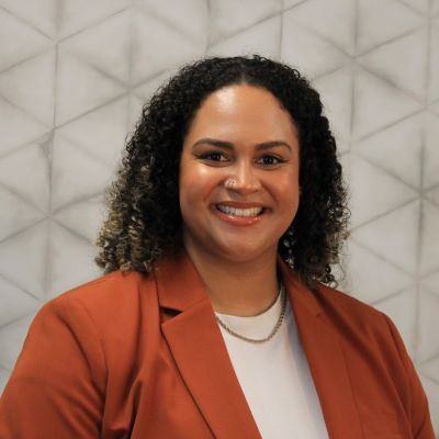 headshot of helena, a black woman, wearing a rusty orange jacket over a white shirt and smiling at the camera. Her hair is shoulder length and curly.