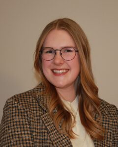 Megan wearing a small black and tan checkered suitjacket smiling at the camera. She is wearing glasses and her hair is down by her shoulders. She is in front of a beige background.