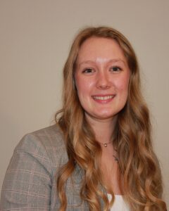 Photo of Sky in a light plaid suitcoat smiling at the camera. Her hair is down and slightly curled. She is in front of a beige background.