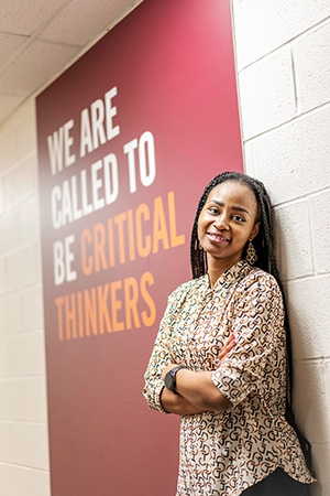Hudani, BIPOC woman standing against wall with hanging behind her that says "We are called to be critical thinkers"