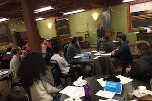 students gathered at tables in a small room listening to someone speak from a stage