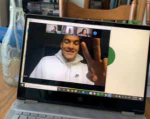 laptop screen with man in white sweatshirt giving peace sign on the screen and three other virtual meeting attendees