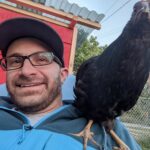 A bearded masculine presenting person waring eyeglasses and a baseball cap, seated in front of a red shed with a chicken perched on his shoulder.