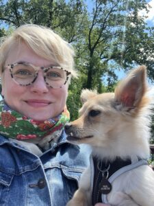 A blonde, middle-aged white woman with short hear, wearing glasses and smiling, with a small blonde dog with pointy ears and a snaggletooth.