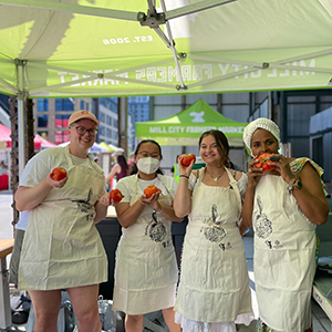 Four people holding tomatoes under a green canopy at a farmers market, wearing aprons.