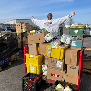 A person stands on a cart full of boxes and containers in a parking lot, smiling with arms outstretched.