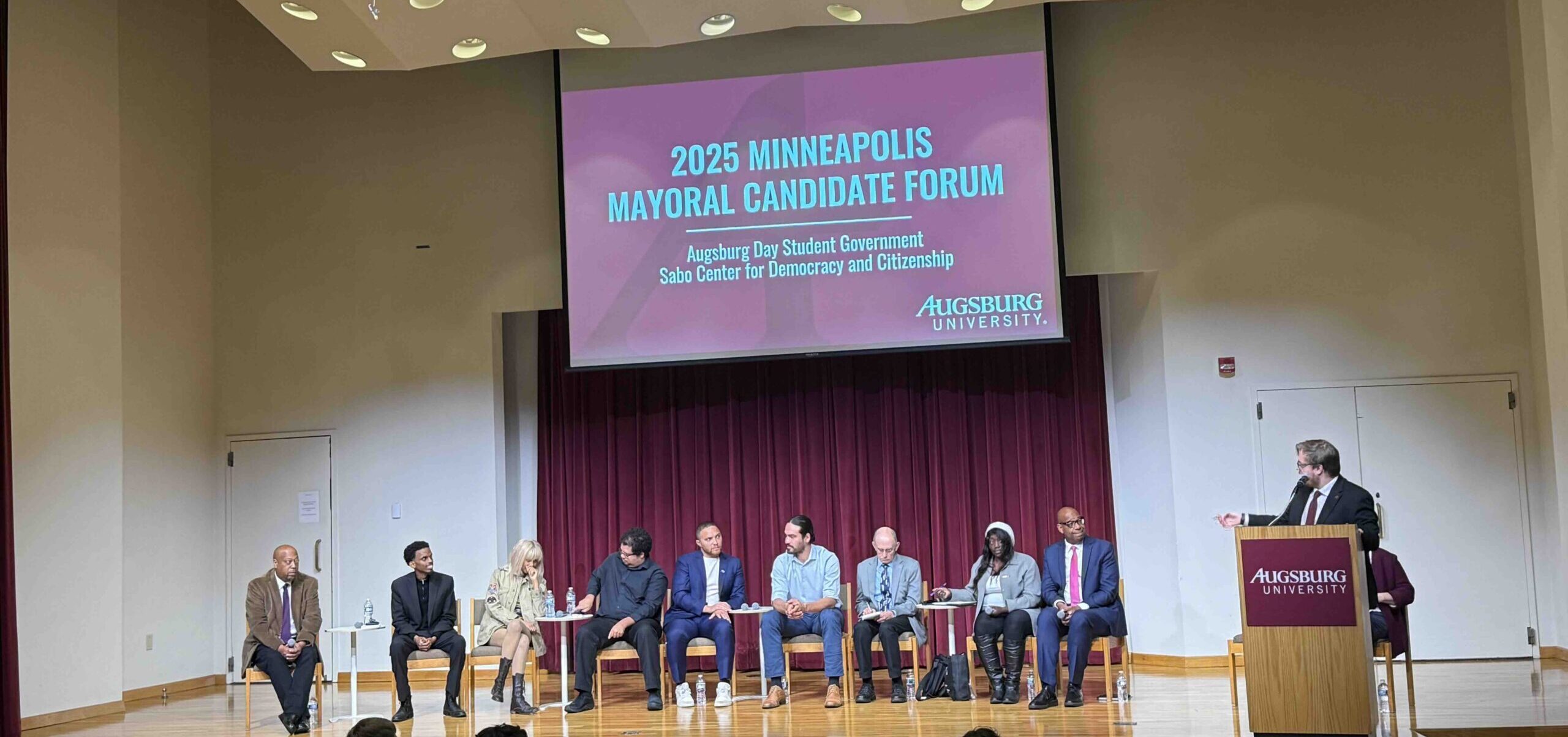 Image of Aidan Wippich leading the Augsburg Mayoral Forum, with 9 candidates on stage.