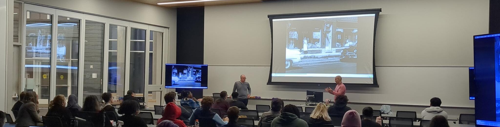 Picture of the movie showing in action, two people at the front of the room talking with the film on the projector