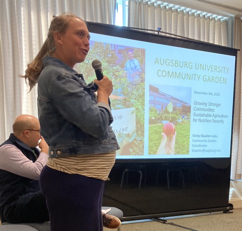 Picture of Greta, with a microphone, standing with others at a panel with slides about the Community Garden in the background.