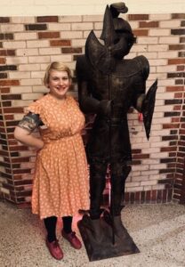 A middle aged woman with short blonde hair, wearing a red dress, stands smiling next to a suit of armor.