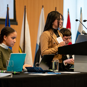 A female student stands at a podium with a laptop, preparing to speak to an audience.