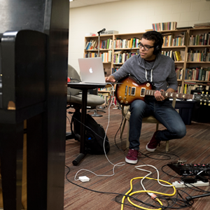 A young man with headphones playing an electric guitar while looking at a laptop in a room with bookshelves and musical equipment.