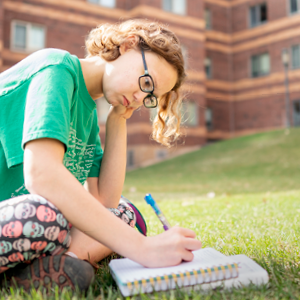 Creative-Arts-Website-Elements11-1.png Image Breakdown Long Description: The image shows a person sitting on a grassy area in front of a red brick building. They are wearing a green t-shirt and glasses, with curly light brown hair. The person is focused on writing in a spiral notebook, holding a pen in hand. The building in the background has multiple stories with uniformly spaced windows. The scene appears to be set on a sunny day, as the grass is brightly lit. Alt-text: Person sitting on grass writing in a notebook outside a red brick building.