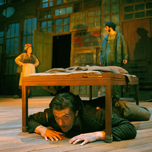 Man lying under a wooden table on stage, with a woman and man standing in the background.