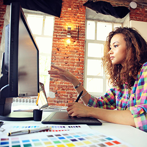 A person using a graphic tablet with a computer, surrounded by a color palette and natural light from windows.