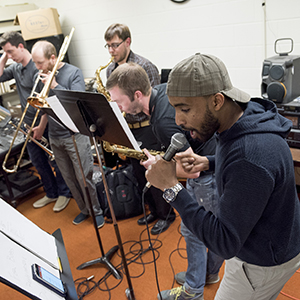 Musicians rehearsing in a practice room, one singing into a microphone with others playing brass instruments.