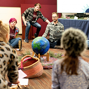 People sitting around a model globe on a wooden floor, with decorative greenery and textiles.