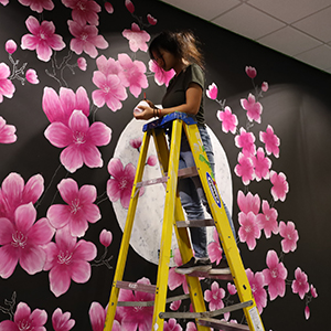 Person on ladder painting pink cherry blossoms on a mural with a moon in the background.